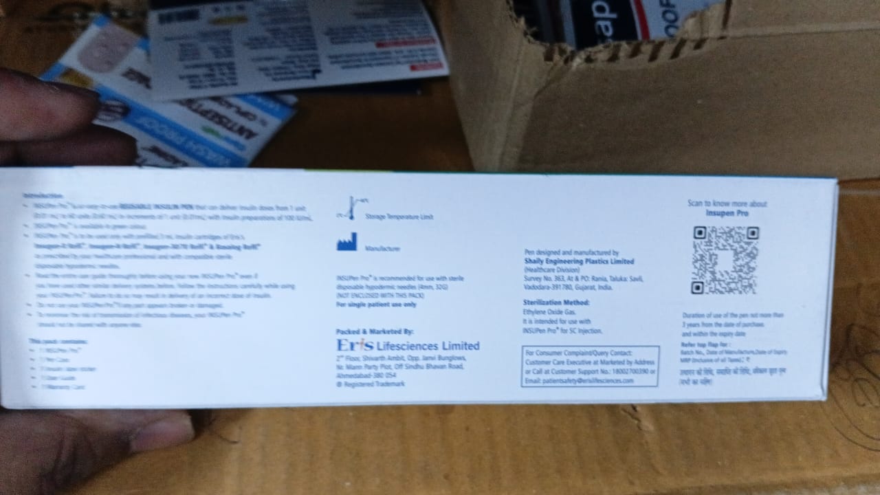 A hand holding the back of a white product box on a cardboard surface, with shipping labels and a box visible in the background.